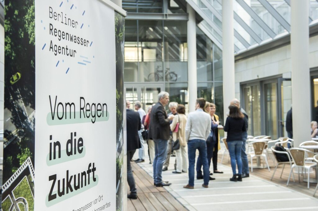 Banner der Veranstaltung Reden über Regen im Vordergrund und im Hintergrund stehende Menschen im Gespräch
