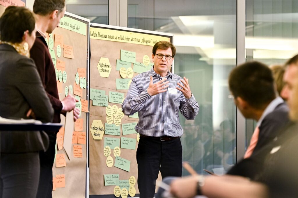 Ein Mann spricht vor einer Pinnwand mit farbigen Zetteln zu umstehenden Teilnehmenden