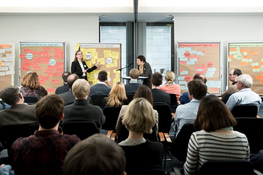 Zwei Personen präsentieren Ergebnisse vor mehreren, in Stuhlreihen sitzenden Personen