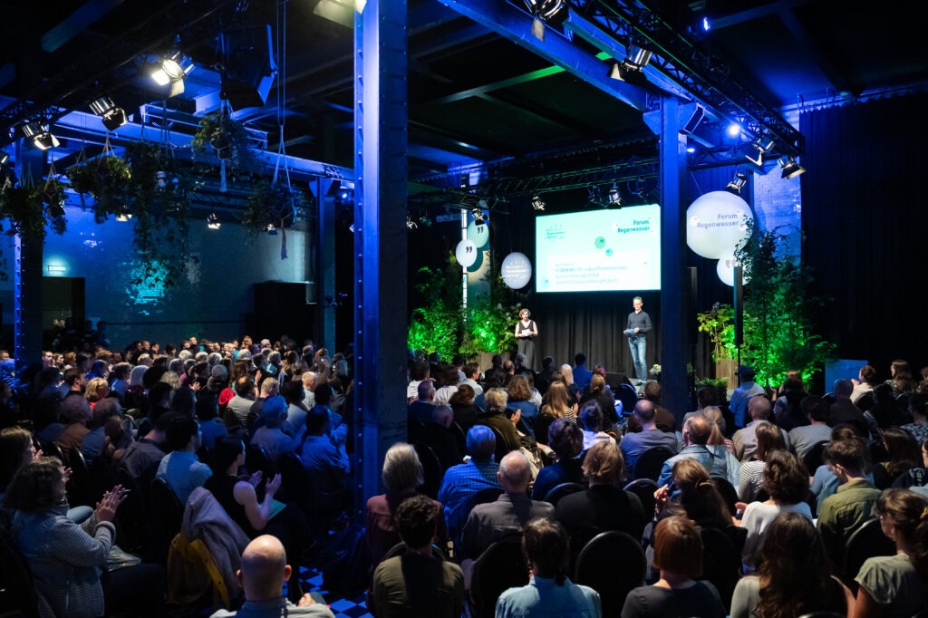 Zwei Personen stehen vor großem Publikum auf der Bühne